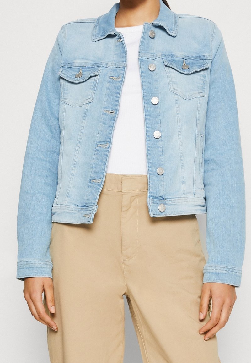 Femme portant une veste en jean bleu clair sur un top blanc et un pantalon beige, debout devant un fond uni.