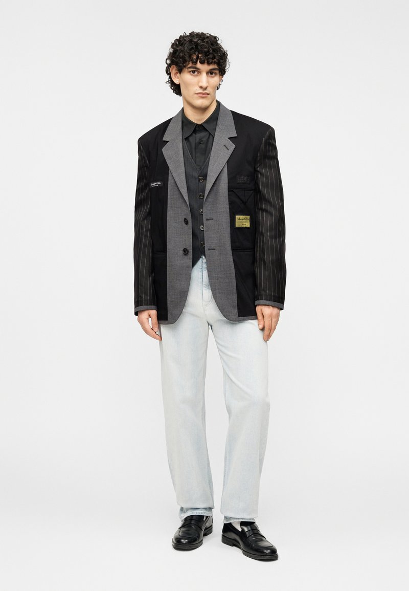 Veste noire et grise avec manches rayées, fermeture à boutons et détails en patch, associée à un jean bleu clair et des chaussures noires.