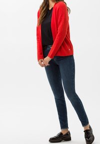 Czerwony sweter robiony na drutach z otwartym przodem i długimi rękawami, zestawiony z ciemnoniebieskimi dopasowanymi dżinsami i czarnymi lakierowanymi butami.