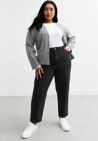 Cardigan gris de punto con cierre de botones, lleva puesto sobre una blusa blanca. La modelo viste jeans negros de tiro alto y zapatillas blancas sobre un fondo neutro.