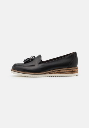 Mocassin en cuir noir avec des glands, semelle en liège superposée contrastée et semelle en caoutchouc blanc, talon bas et design slip-on sur fond clair.