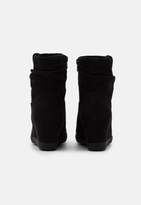 Bottes de cheville en daim noir avec un design froncé, dotées de fermetures éclair latérales et d'un talon compensé pour plus de stabilité. Texture lisse et détails minimalistes.