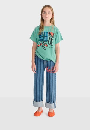 Ragazza in piedi che indossa una maglietta verde con stampa di cane, jeans larghi blu con testo rosso e scarpe slip-on arancioni su sfondo bianco.