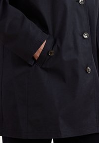 Ναυτικό μπλε αδιάβροχο παλτό με κουμπωμένη μπροστινή πλευρά, πλαϊνές τσέπες και υφασμάτινες μανσέτες. Διαθέτει μεγάλες μαύρες κουμπωτές για κλείσιμο.