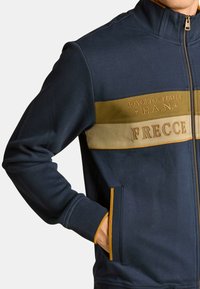 Chaqueta con cremallera en color marino con rayas horizontales en oro y verde olivo. Cuenta con puños acanalados, bolsillos laterales y texto bordado en el pecho.