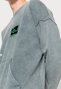 Szara bluza z dużą kieszenią z przodu, prążkowanymi mankietami, ozdobną haftowaną naszywką i teksturowanym wykończeniem.