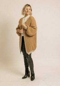 Cardigan en tricot beige à coupe ample, texture côtelée et poignets en gris clair contrastant. Associé à un pantalon noir ajusté et des bottes à talons.