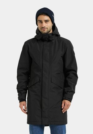 Mand iført en sort hættejakke til vinter med klaplommer og en mørkeblå strikket hue med blå jeans.