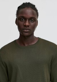 Olijf groene gebreide trui met een ronde halslijn, gestructureerde stof en lange mouwen. Behalve subtiele ribbeldetails langs de kraag en manchetten.