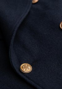 Marineblauer Wollstoff mit glatter Textur, versehen mit einem goldenen Knopf mit einem kunstvollen Emblemdesign und abgerandetem Rand.