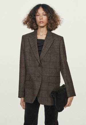 Blazer a quadri marroni con rever a punta, chiusura con bottone anteriore e due tasche frontali, abbinato a un top scuro e con una pochette in mano.