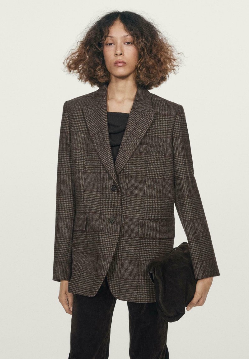 Blazer a quadri marroni con rever a punta, chiusura con bottone anteriore e due tasche frontali, abbinato a un top scuro e con una pochette in mano.
