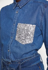 Denim shirt with a button-down collar, featuring a silver sequin pocket. The fabric is blue, with a belted waist and classic fit.