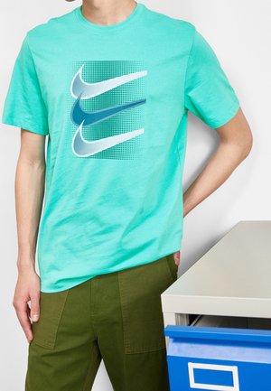 Persona che indossa una maglietta turchese con tre loghi Nike Swoosh sovrapposti, in piedi accanto a una scrivania bianca con un cassetto blu parzialmente aperto.