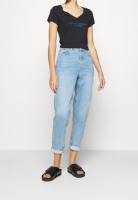 Black V-neck t-shirt with "CALVIN KLEIN JEANS NEW YORK" in turquoise, light blue high-waisted jeans, and black slide sandals.