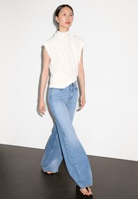 White sleeveless top with draped collar, paired with light blue flared jeans. Smooth fabric, casual fit, and open-toe footwear.