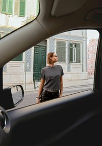 Mladá žena v tmavém oblečení stojí poblíž vzorované evropské budovy, pozorována skrze okno auta na městské ulici.