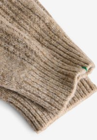 Vêtement en laine beige tricoté présentant une texture côtelée avec un ourlet replié. Un petit accent vert est visible sur un bord.