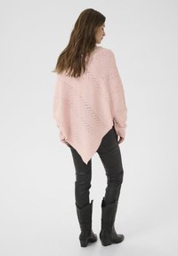 Ženska obrnjena s strani, oblečena v roza pleteno pončo, črne usnjene hlače in črne kavbojske čevlje, na enobarvni svetli podlagi.