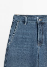 Blue denim jeans waist area showing belt loops, metal button, front pockets, and visible stitching on a white background.