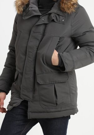 Person trägt eine dunkelgraue Winterjacke mit pelzbesetzter Kapuze, Hand in der Seitentasche und dunkle Jeans vor weißem Hintergrund.