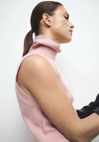 Svijetlo ružičasti džemper bez rukava s rol-kragnom i rebrastom teksturom, s širokim otvaranjima za ruke i detaljima razdvajanja sa strane. Minimalistički dizajn, mekani materijal.