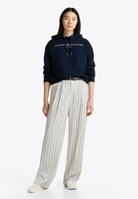 Granatowy hoodie z białym logo i sznurkami, w połączeniu z białymi spodniami w szerokim kroju z kolorowymi pionowymi paskami. Całość dopełniają białe trampki.