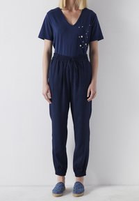 Navy blauwe jumpsuit met een V-hals, korte mouwen, elastische taille en taps toelopende benen. Decoratieve knopen op de borstzak.