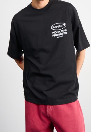 Person iført en sort Carhartt Work In Progress T-shirt med hvidt logo og røde bukser, stående med hænderne bag ryggen.