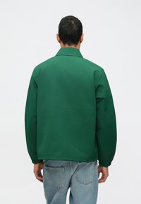 Πράσινο σακάκι με γιακά, φτιαγμένο από μαλακό ύφασμα; διαθέτει ελαστικούς μανσέτες και χαλαρή γραμμή; συνδυασμένο με ανοιχτόχρωμα μπλε τζιν.