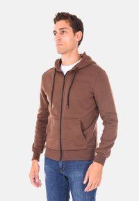 Felpa con zip marrone realizzata in un tessuto morbido, dotata di tasca frontale, cappuccio regolabile con cordino e polsini a coste. Indossata sopra una t-shirt bianca.