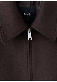 Veste en laine marron foncé avec col pointu et tirette de fermeture éclair argentée, montrant une étiquette noire avec texte blanc "MNG" à l'intérieur du col.