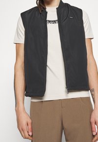 Černá bezrukávová vesta vyrobená z hladkého, lehkého materiálu se zipem vpředu a diskrétním logem na hrudi.