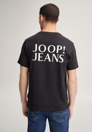 Homme de dos, portant un t-shirt noir avec un grand texte blanc "JOOP! JEANS" et un jean bleu, debout devant un fond clair uni.