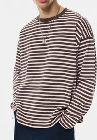 Osoba w brązowej i jasnoróżowej poziomo paskowanej koszuli z długim rękawem, z srebrnym wisiorkiem na szyi, ręce w kieszeniach ciemnoniebieskich spodni.