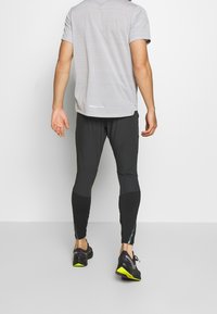 Camisa gris de manga corta, leggings negros ajustados con paneles texturizados y zapatillas deportivas negras con acentos amarillos. Vista desde atrás.