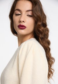 Pulover tricotat crem, cu un finisaj texturat, având mâneci bufante și un decolteu rotund. Modelul are părul ondulat maro și ruj de culoare închisă.