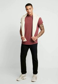 Jonge man met kort haar staat in een bordeauxrood t-shirt, zwarte broek, witte sneakers en een licht sweater over zijn schouders gedrapeerd.