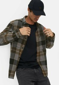 Koszula flanelowa w odcieniu szarym z brązowym, czarnym i białym wzorem w kratę, zapinana na guziki z przodu, a pod nią czarny t-shirt z długim rękawem. Na głowie czarna czapka.
