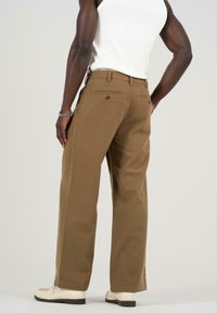 Pantalones de pierna ancha marrones hechos de una tela texturada, con bolsillos traseros y un detalle de botón. Llevados con una camiseta sin mangas blanca y zapatos.