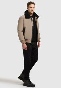 Beige bomberjakke med svart kunstpelskrage, glidelåslommer, ribbestrikkede mansjetter, brukt over en svart skjorte og svarte bukser, kombinert med svarte sko.