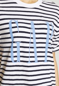 Biała i granatowa koszulka w paski z krótkimi rękawami. Z przodu duża niebieska grafika "GAP" na środku. Wykonana z miękkiego materiału bawełnianego.