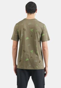 Camiseta de algodón verde oliva con varios logotipos de Nike en negro y verde brillante sobre la tela, con un ajuste relajado y mangas cortas.