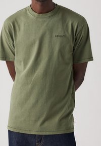 Persona vestida con camiseta olive green de Levi’s y vaqueros azul oscuro, de pie con las manos detrás de la espalda frente a un fondo liso.