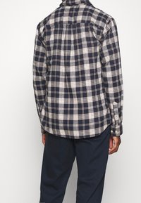 Koszula w kratę w odcieniach granatu, beżu i kremu. Posiada zapięcia na guziki przy mankietach oraz zaokrąglony dół. Wykonana z miękkiego materiału o teksturowanym wykończeniu.