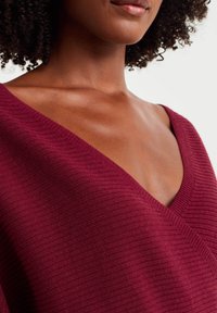 Bordeauxroter V-Ausschnitt-Pullover mit gerippter Textur. Verfügt über einen tiefen Ausschnitt und weichen Stoff, der eine körpernahe Silhouette und ein dezentes Muster betont.