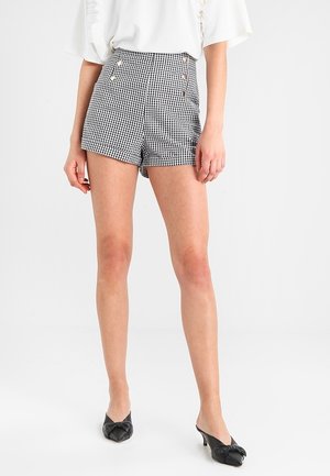 Shorts à carreaux noir et blanc avec taille haute, dotés de deux boutons dorés et d'une coupe ajustée, associés à des chaussures noires.