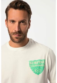Bílý bavlněný tričko s grafickým motivem zeleného opotřebovaného štítu a textem "SURFING CALIFORNIA" spolu s designem zkřížených pádla na levé straně hrudi.