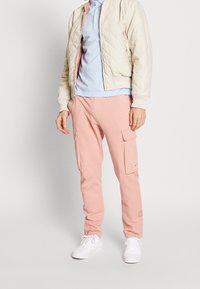 Ljusrosa joggerbyxor med två cargofickor, i kombination med en himmelblå polo och en krämfärgad, vadderad bomberjacka. Vita sneakers fulländar looken.