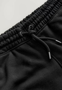 Schwarze Baumwoll-Sweatpants verfügen über einen elastischen Taillenbund mit geraffter Textur und einem Kordelzug mit metallischen Aglets. Glattes Material, ohne Muster.